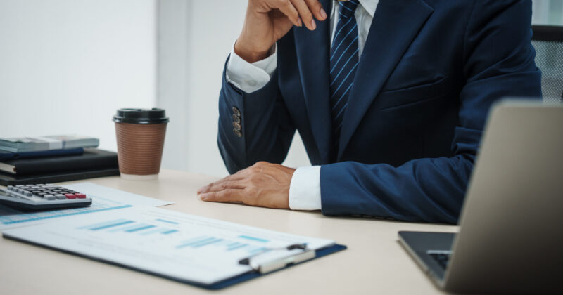 A banker in a suit works at a desk handling loans, analyzing client financial data, providing financial advice, dealing with stocks and the stock market, and studying economic situations. Business professional seated at a desk reviewing financial documents beside a calculator, coffee cup, and laptop.